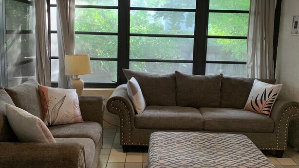 A living room with a sofa, armchair, ottoman, and a lamp. There are large windows with curtains, and a ceiling light fixture.