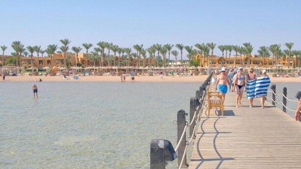 A wooden pier extending into a clear beach with palm trees and a row of buildings in the background.