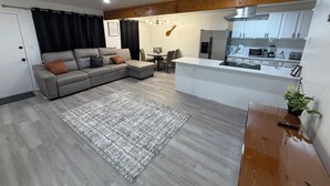 A modern living room with a grey sectional sofa, a small dining table, and a kitchen area with white cabinets and appliances.
