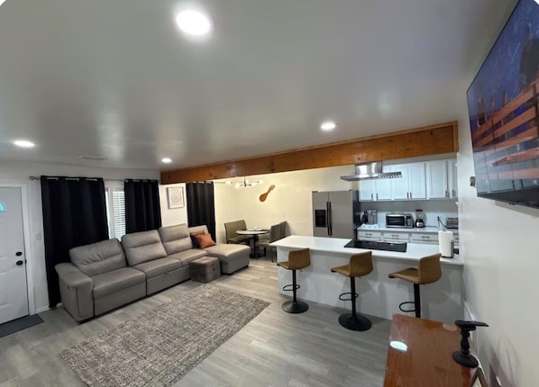 A modern living room with a sofa, ottoman, and a coffee table. There is a kitchen area with a white countertop and bar stools. - Charming 3-bedroom with WiFi and AC in Midwest City (Midwest City)