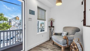 A room with a balcony, a wooden floor, a chair, a vase with flowers, and a hanging light fixture. - Professionally Managed | Walk to Beach (Seaside Heights)