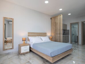 A modern bedroom with a wooden bed frame, a nightstand with a lamp, a large mirror, and a bathroom visible through a partition. - Πολυτελής σουιτα με ιδιωτικη πισινα και υπέροχη θέα στο Αιγαίο! (Paros)