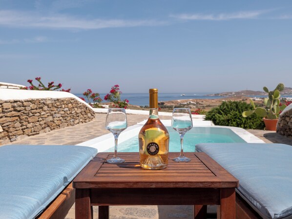 A wooden table with a bottle and two glasses, overlooking a pool and a scenic view of the sea and hills. - Πολυτελής σουιτα με ιδιωτικη πισινα και υπέροχη θέα στο Αιγαίο! (Paros)