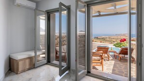 A modern room with a sliding glass door leading to an outdoor seating area with a pool, stone walls, and a view of the sea. - Πολυτελής σουιτα με ιδιωτικη πισινα και υπέροχη θέα στο Αιγαίο! (Paros)