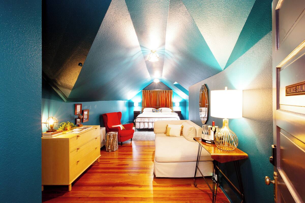 Camera da letto con soffitto spiovente, un letto, una sedia rossa, un divano bianco e un comò in legno.