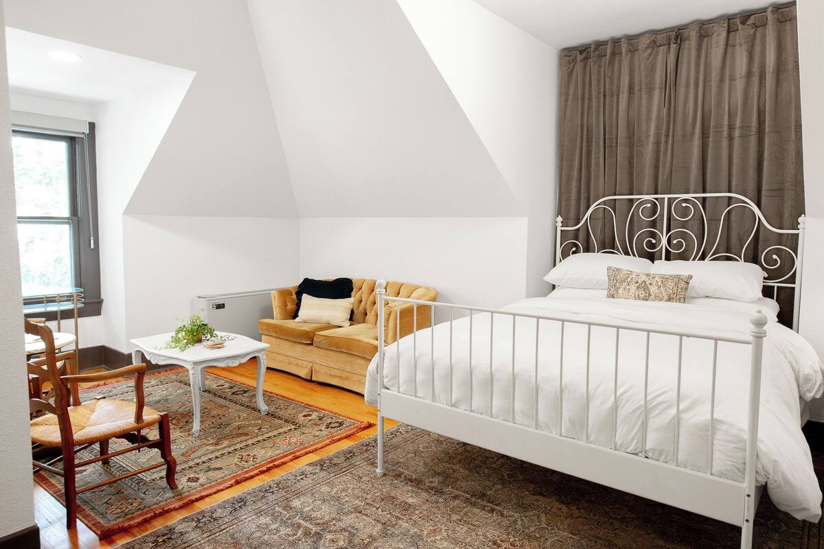 Camera da letto con un letto in ferro bianco, una sedia in legno, un tavolino bianco con una pianta, un divano beige e un tappeto a motivi.