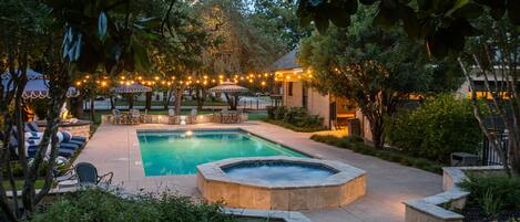 Una piscina con hidromasaje, rodeada de sillas de descanso y sombrillas, con luces colgantes.