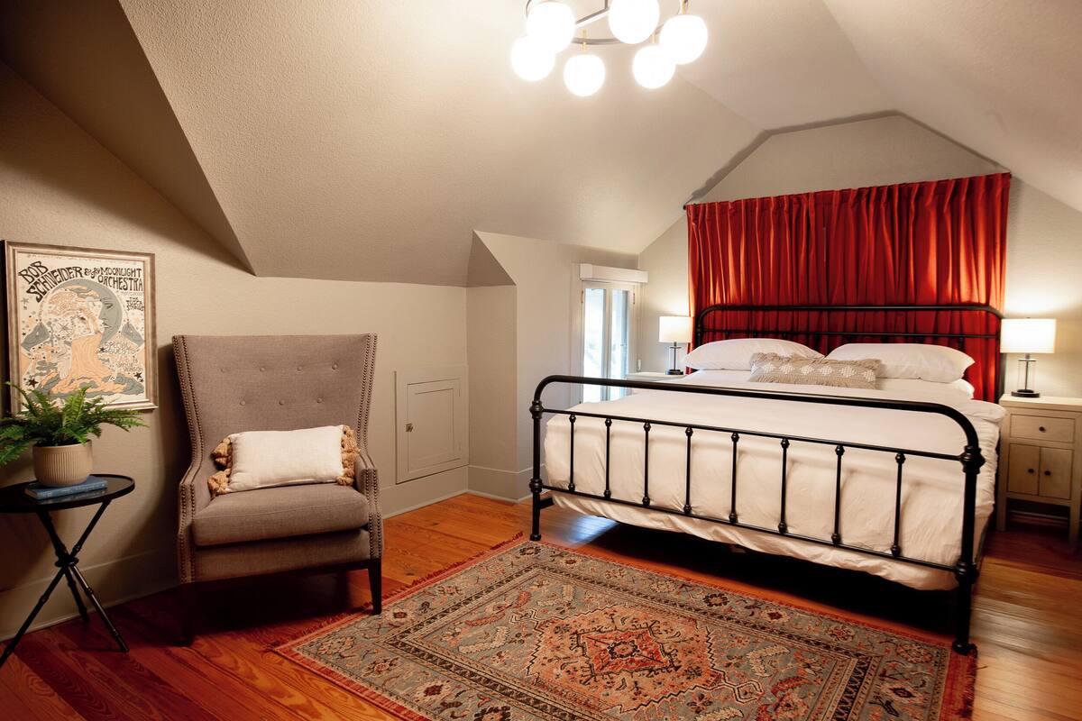 Una camera da letto con un letto, una sedia, un comodino, una lampada, un tappeto e un quadro appeso al muro.