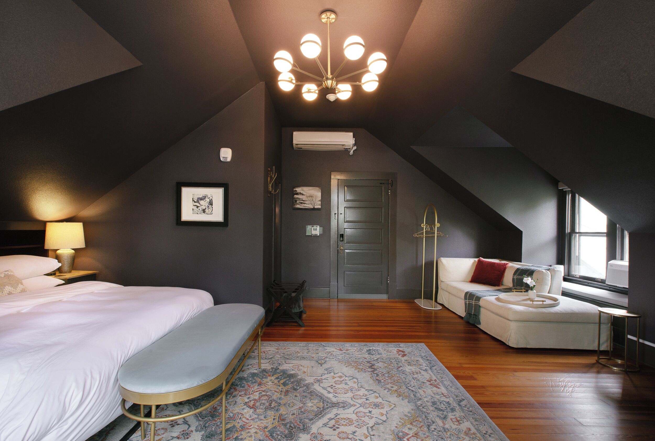 A modern bedroom with a large bed, a bench, a sofa, a chandelier, and a view of the outdoors.