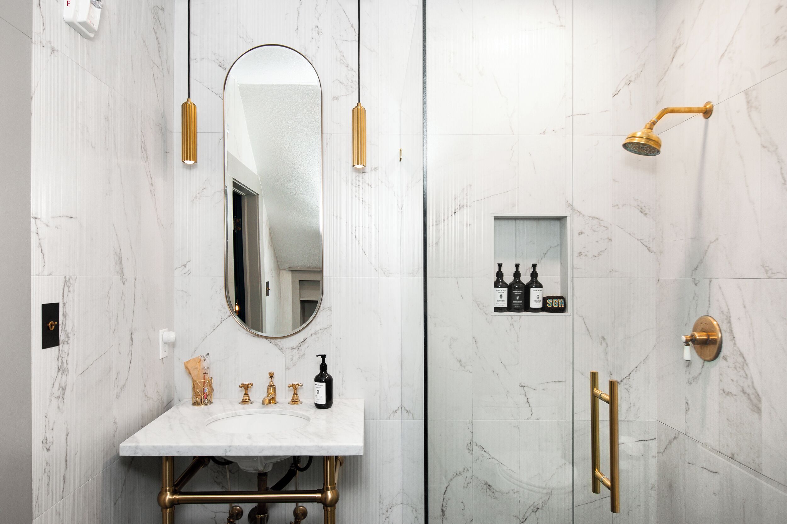 Un baño con pared de mármol, lavamanos con marco dorado, un espejo grande, cabezal de ducha dorado y un estante pequeño con artículos de tocador.