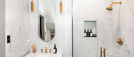 Un baño con pared de mármol, lavamanos con marco dorado, un espejo grande, cabezal de ducha dorado y un estante pequeño con artículos de tocador.