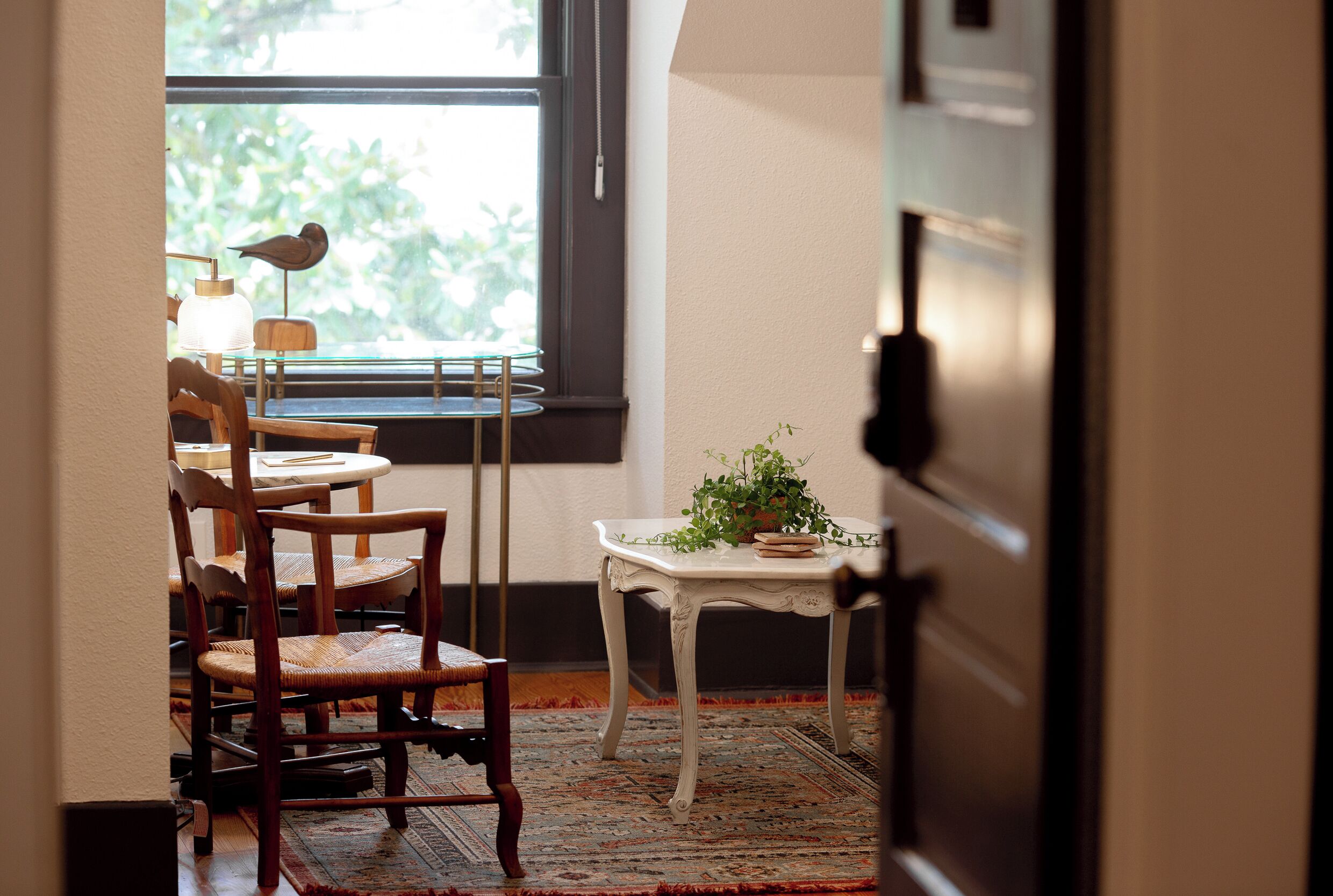 Una habitación con comedor que incluye sillas de madera y una mesa, una pequeña mesa blanca con una planta, y una ventana con vistas a zonas verdes.