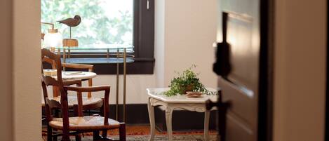 Una habitación con comedor que incluye sillas de madera y una mesa, una pequeña mesa blanca con una planta, y una ventana con vistas a zonas verdes.