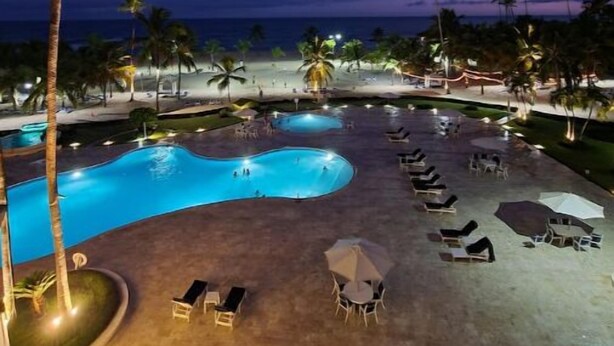 A resort pool area with lounge chairs and umbrellas, palm trees, and a view of the ocean at dusk.