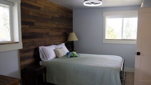 A bedroom with a wooden headboard, a bed with a green bedspread, a nightstand, a lamp, and a window with a view of trees. - The Cottage at Dragonfly Meadows Farm with AC and wifi (Cheney)