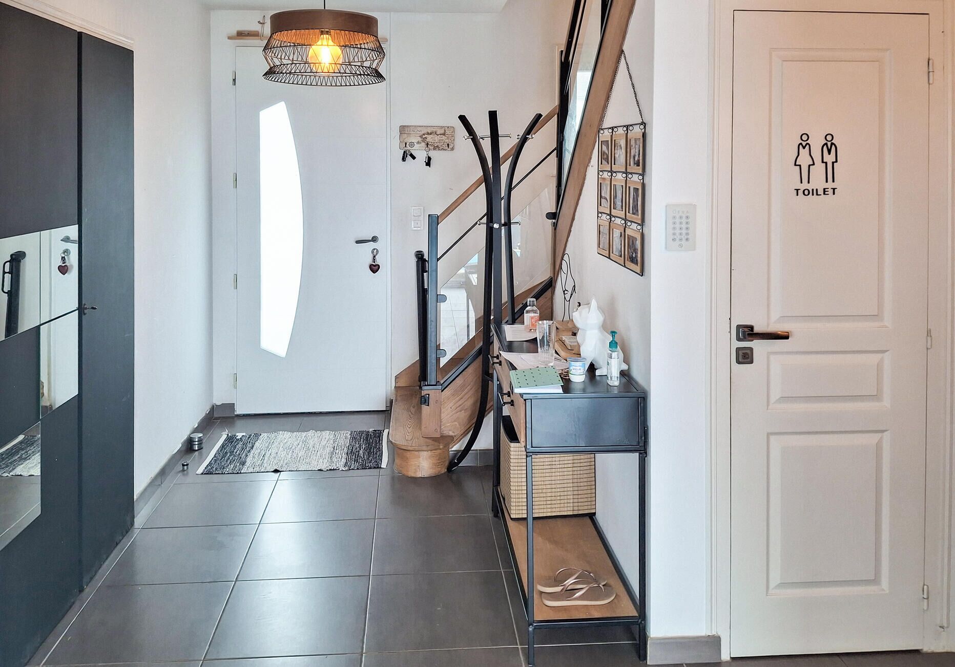 Een moderne hal met een trap, een witte deur met een toiletpictogram en een klein tafeltje met persoonlijke spullen.