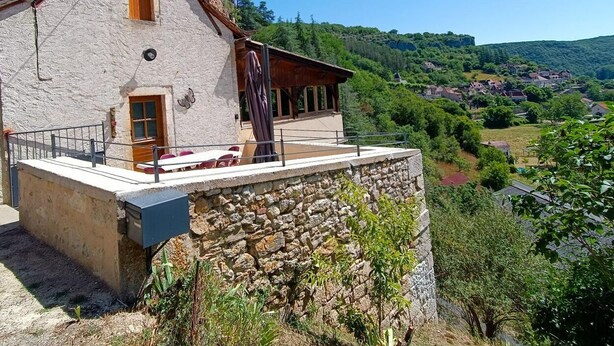 A stone building with a wooden door and window, a balcony with a table and chairs, and a view of a valley with houses. - Gîtes de France® - L'Oustal de Marguerite (SAINT SULPICE)
