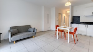 A modern living room with a grey sofa, a dining table with orange chairs, and a kitchen area with white cabinets and a black countertop. - Appartement Proche Plage (La Rochelle)