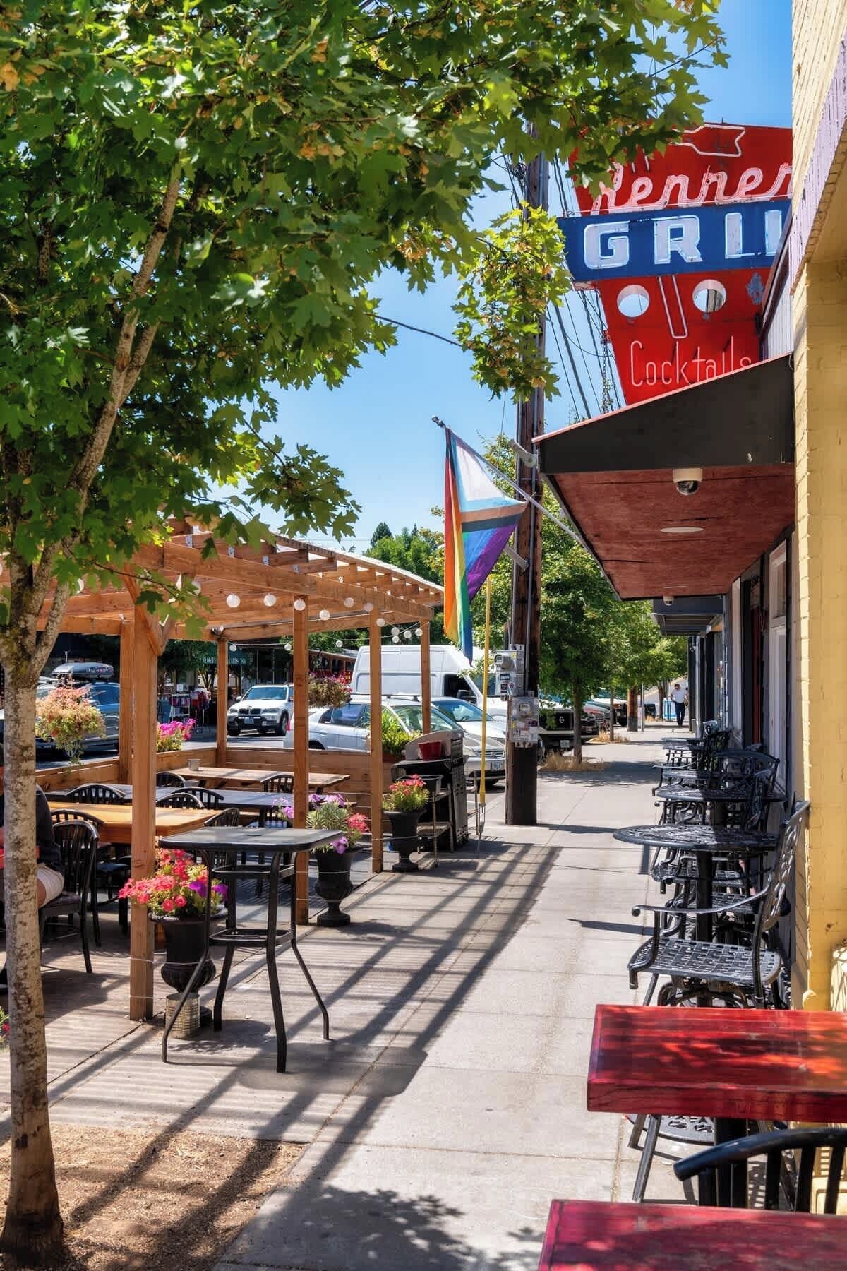 Ein Außenbereich zum Sitzen mit Tischen und Stühlen, eine Holzpergola und ein Gebäude mit einer Neonreklame für Grillgerichte und Cocktails.