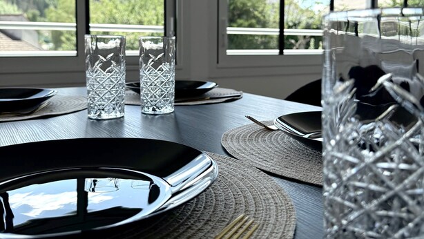 Une table à manger dressée avec des assiettes, des couverts et des verres, donnant sur une forêt grâce à de grandes fenêtres.