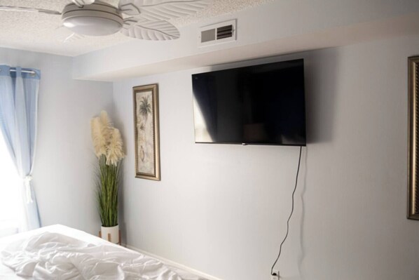 A bedroom with a bed, a wall-mounted TV, a ceiling fan, and a window with curtains.