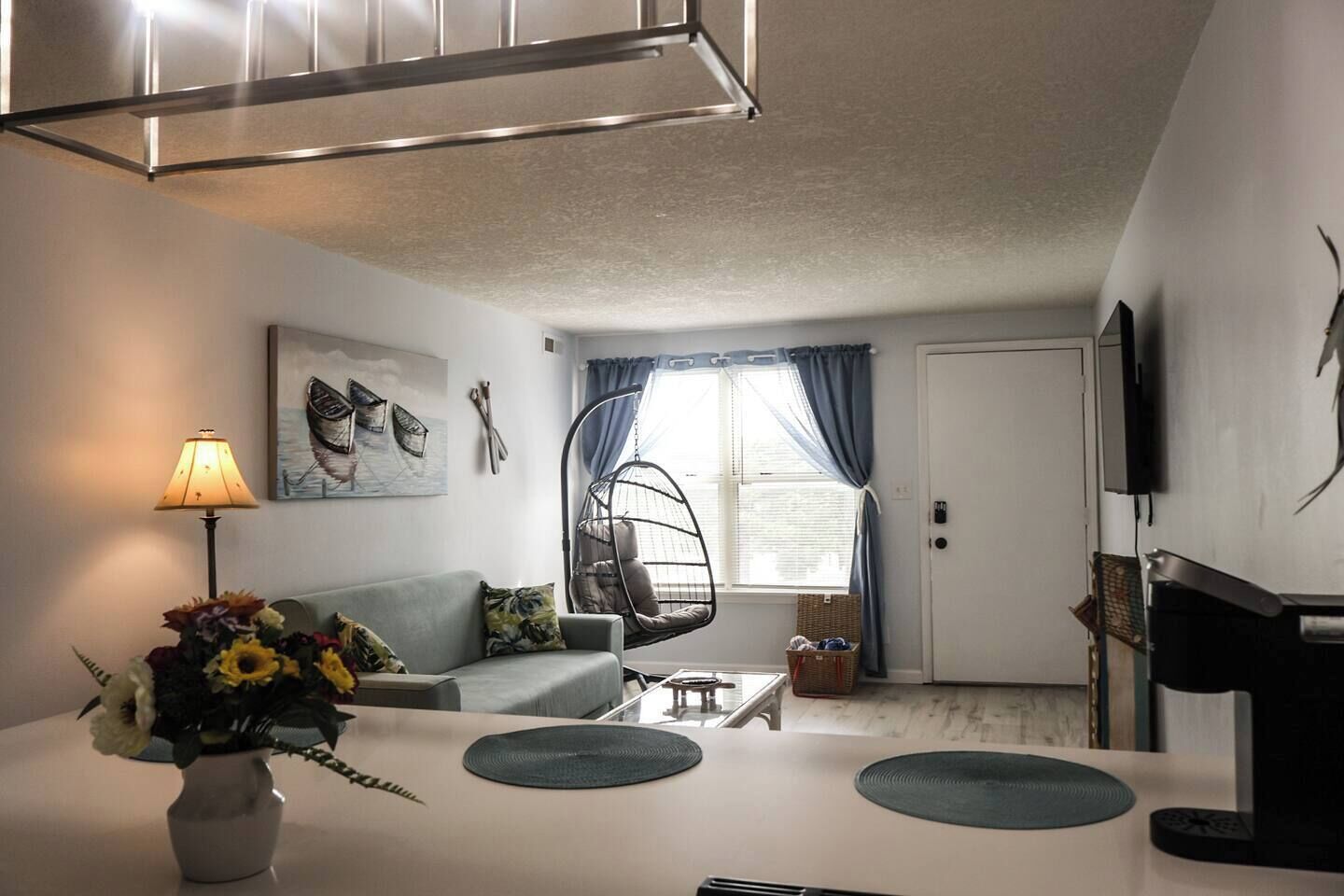 Una sala de estar con un sofá, una silla colgante, una mesa con un jarrón de flores y un televisor en la pared.