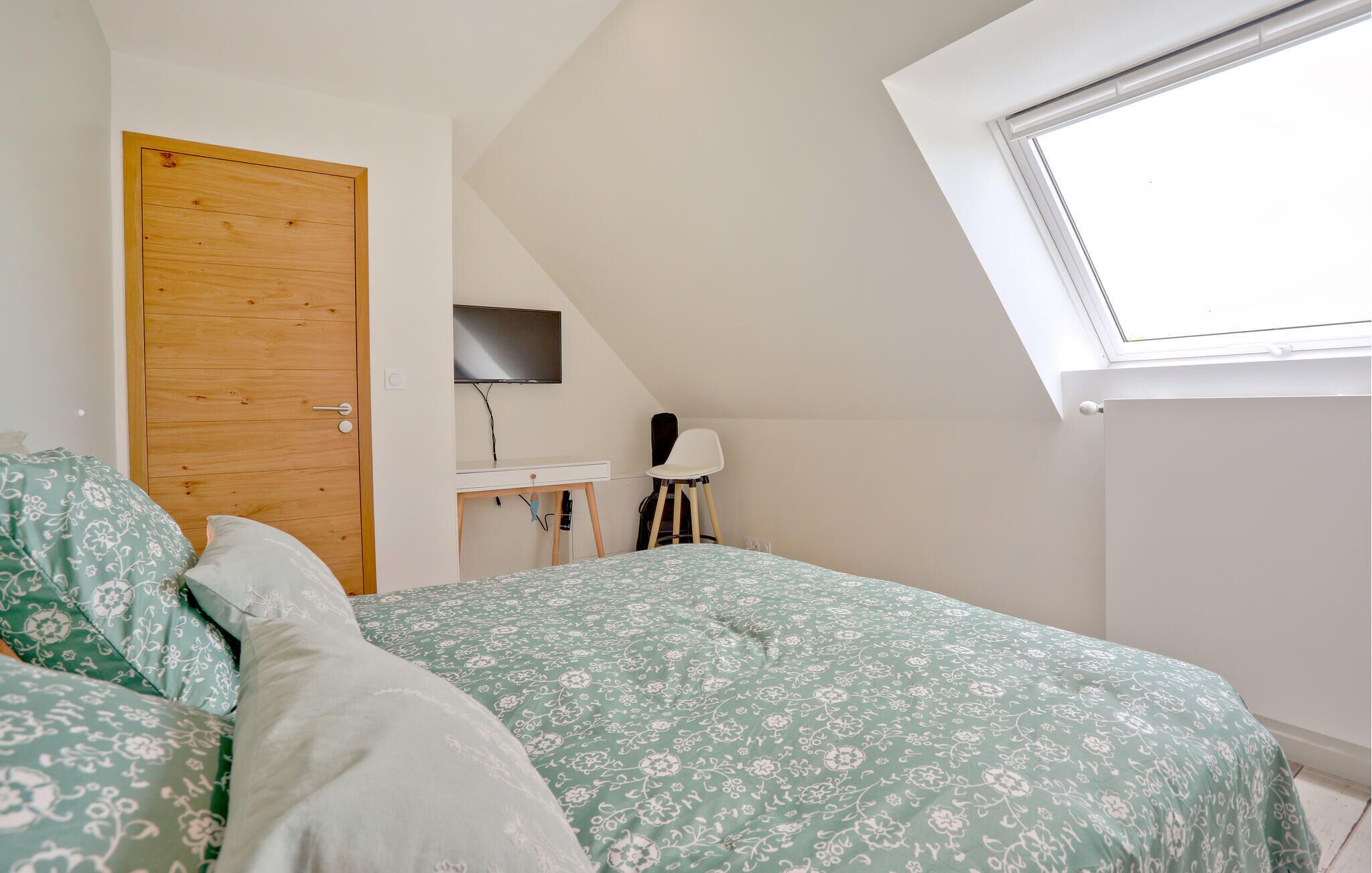 A bedroom with a sloped ceiling, a wooden door, a desk with a chair, a bed with floral bedding, and a skylight.