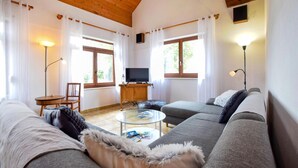 Una sala moderna con un sofá gris, una mesa de centro y un televisor sobre un mueble de madera. Hay dos ventanas con cortinas, una lámpara de pie y una pequeña mesa con una silla.