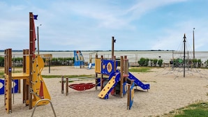 Un parque infantil con diversas estructuras de juego, como toboganes y juegos de escalada, ubicado sobre una zona arenosa cerca de una masa de agua.