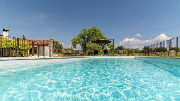 Ein klarer Pool mit einer gefliesten Liegefläche, umgeben von einem Zaun und beschattet von Sonnenschirmen.