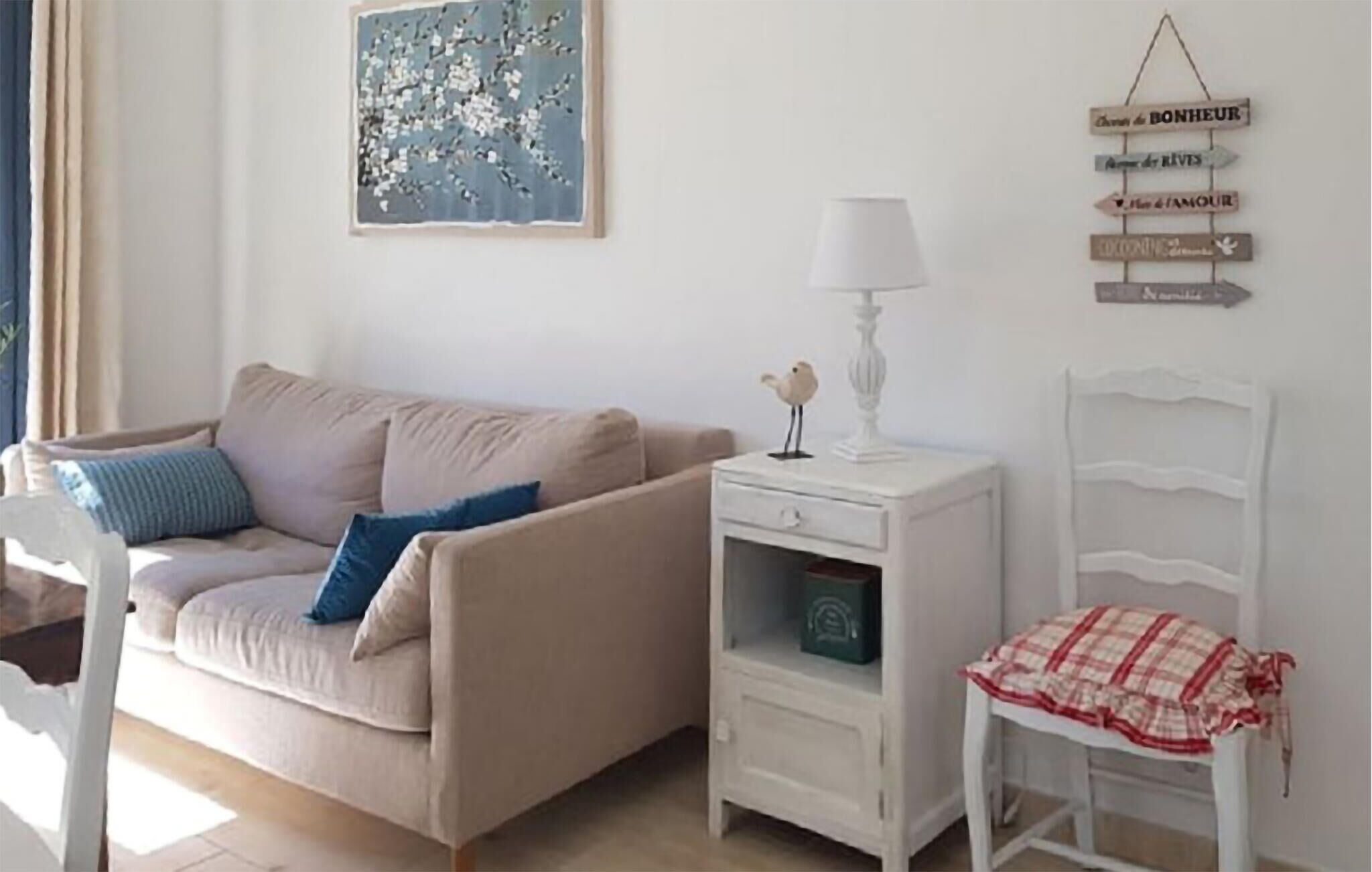 Een woonkamer met een beige bank, een witte bijzettafel met een lamp, een witte stoel met een rood-wit geruit deksel, en een muur met een blauw abstract schilderij en een bord met Franse woorden.