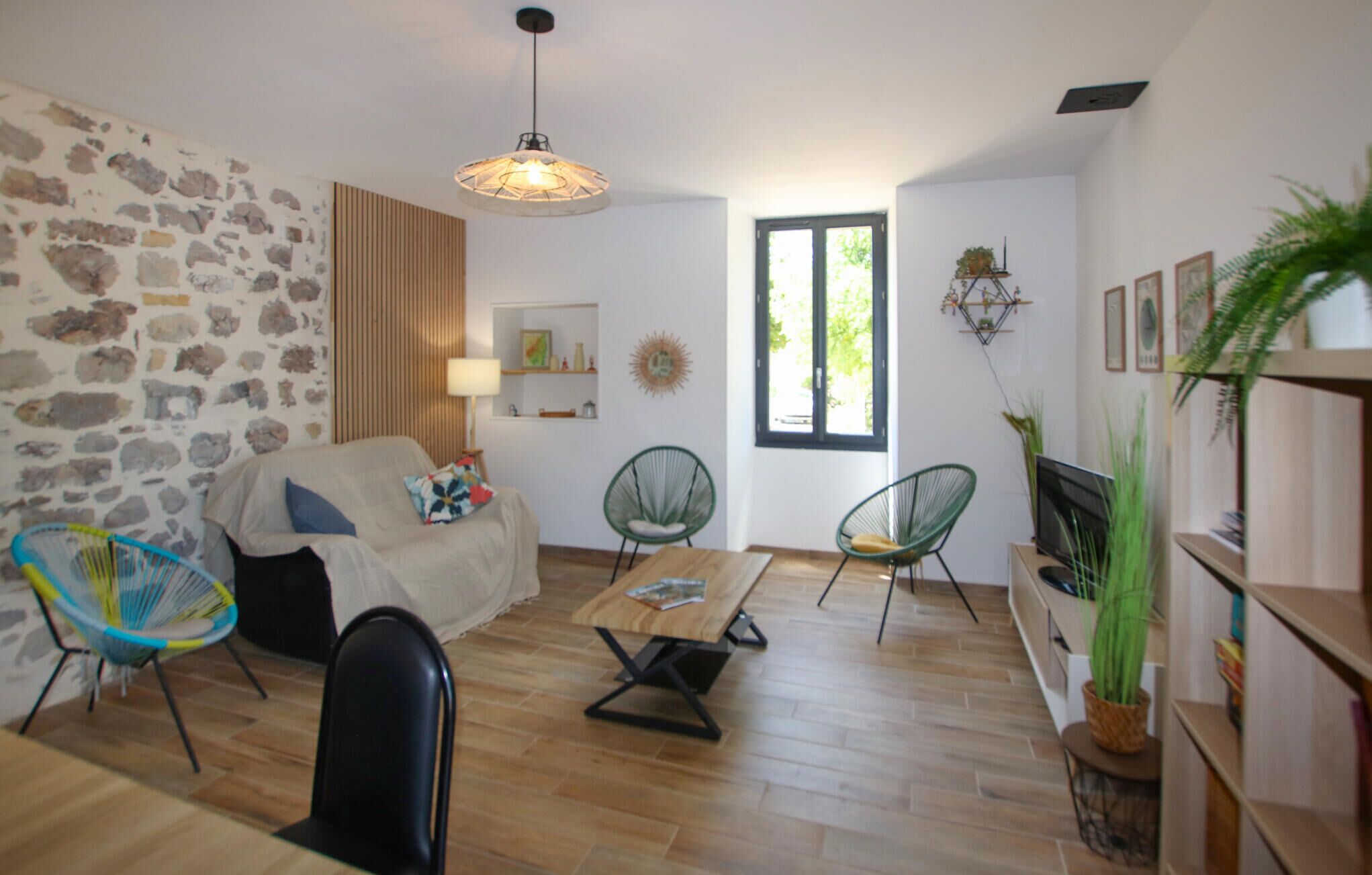 Una sala de estar con pared de piedra, piso de madera, un sofá, dos sillones verdes, una mesa de centro y un televisor.