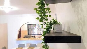 Een moderne eetruimte met een witte tafel en grijze stoelen, een zwart zwevend schap met een plant, en een zicht op de keuken op de achtergrond.