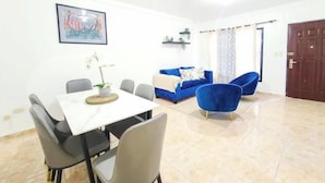 Een eethoek met een tafel en stoelen, een blauwe bank en een blauwe fauteuil in een goed verlichte kamer met witte muren en een betegelde vloer.