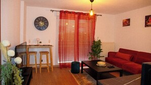 Una sala de estar con un sofá rojo, una mesa de centro de madera, una mesa de comedor con sillas, un reloj en la pared y una cortina roja.