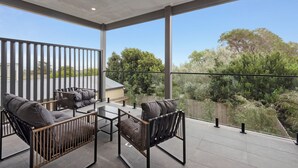 Ein großzügiger Balkon mit modernem Design, ausgestattet mit Glasgeländer, einer Sitzecke mit zwei Sofas und einem Couchtisch, und Blick auf einen üppigen Garten sowie entfernte Gebäude.