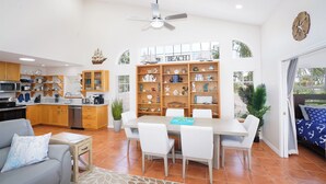 A spacious living area with a dining table, white chairs, a gray sofa, and a kitchen area with wooden cabinets and a ship-themed bookshelf. - 4/3 Condo Block From Beach With Golf Cart, Hot Tub, Kayaks, Games (Indian Rocks Beach)