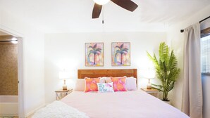 Een slaapkamer met een houten hoofdbord, twee aan de muur bevestigde schilderijen, een potplant en een zachte witte plaid.