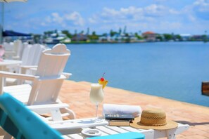 Un área de descanso frente a la playa con sillones blancos, una bebida con cereza en el borde y un sombrero de paja sobre la mesa.