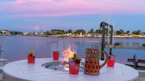 Una mesa con bebidas y un pozo de fuego, con vistas a un cuerpo de agua y edificios al fondo.