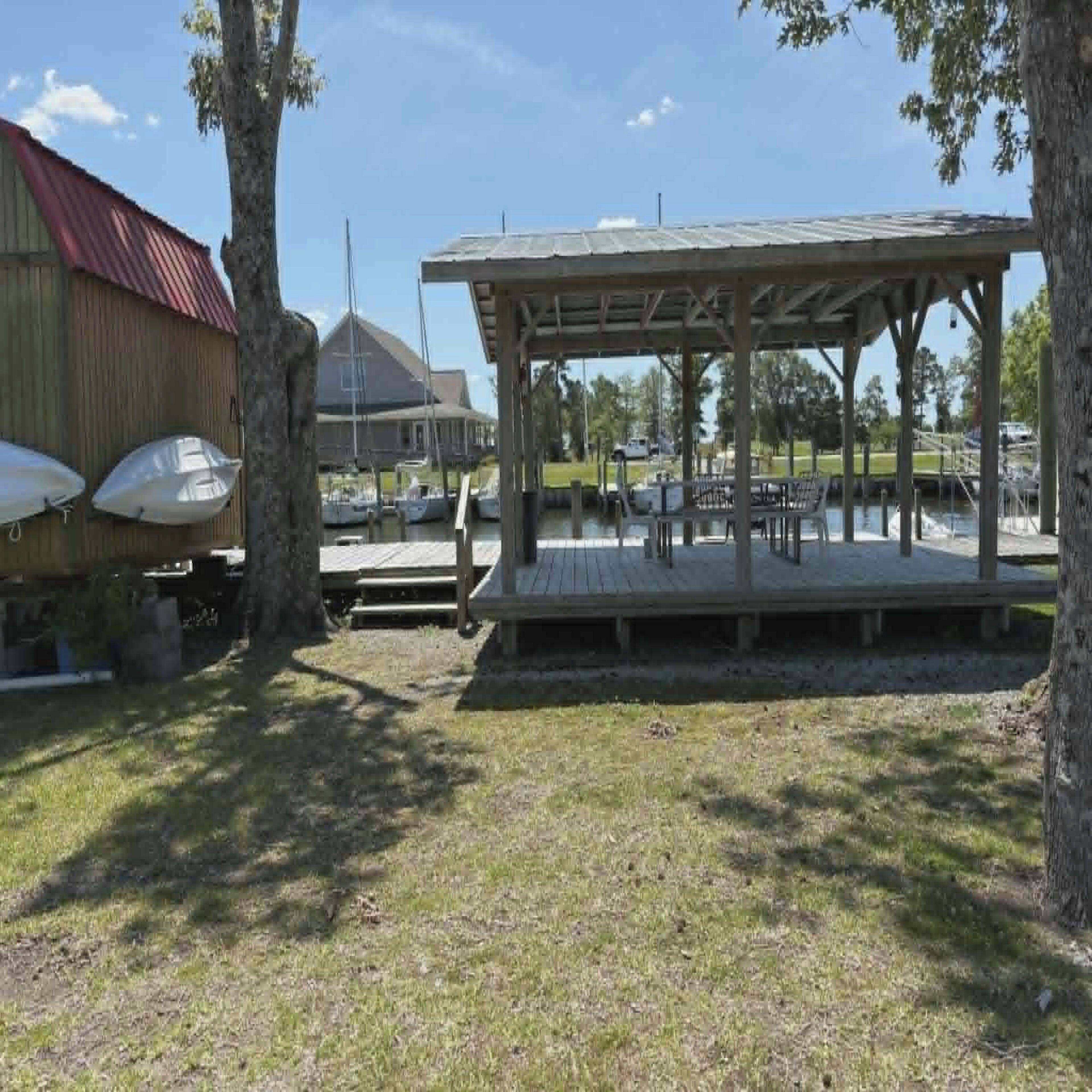 A covered outdoor seating area with a wooden deck, surrounded by trees and a grassy area.