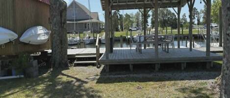 A covered outdoor seating area with a wooden deck, surrounded by trees and a grassy area.