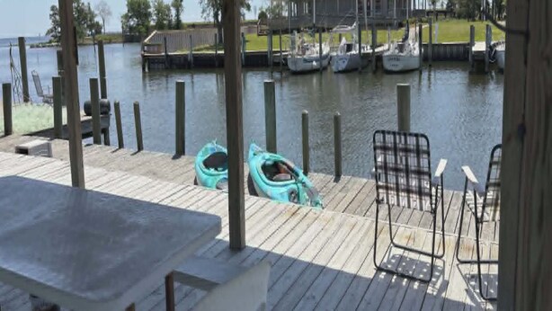 A wooden deck with a table and chairs, a covered area, and a view of a house and boats on the water.