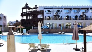 A swimming pool with lounge chairs and umbrellas, surrounded by a white building with balconies and a wooden structure.