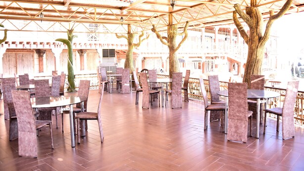An outdoor dining area with round tables and chairs, a wooden ceiling structure, and a view of buildings in the background.