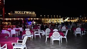 An outdoor seating area with white chairs and tables, a 'HOLLYWOOD' sign in the background, and a fountain with lights.