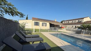A backyard with a swimming pool, two lounge chairs, a small garden, and a white fence.