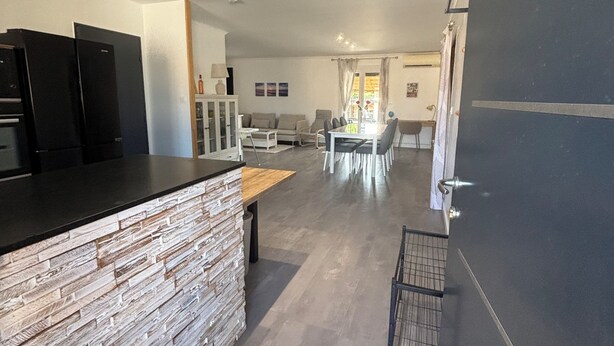 A modern kitchen with a stone backsplash, a dining area with a table and chairs, and a living room with a sofa and a TV.