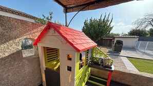 A children's playhouse with a red roof and a green door, situated on a wooden deck with a metal fence in the background.