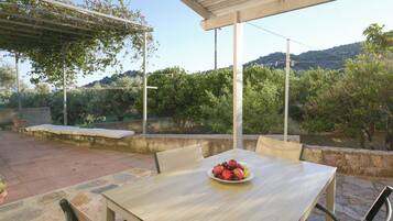 An outdoor patio with a table and chairs, a fruit bowl on the table, and a pergola with climbing plants.